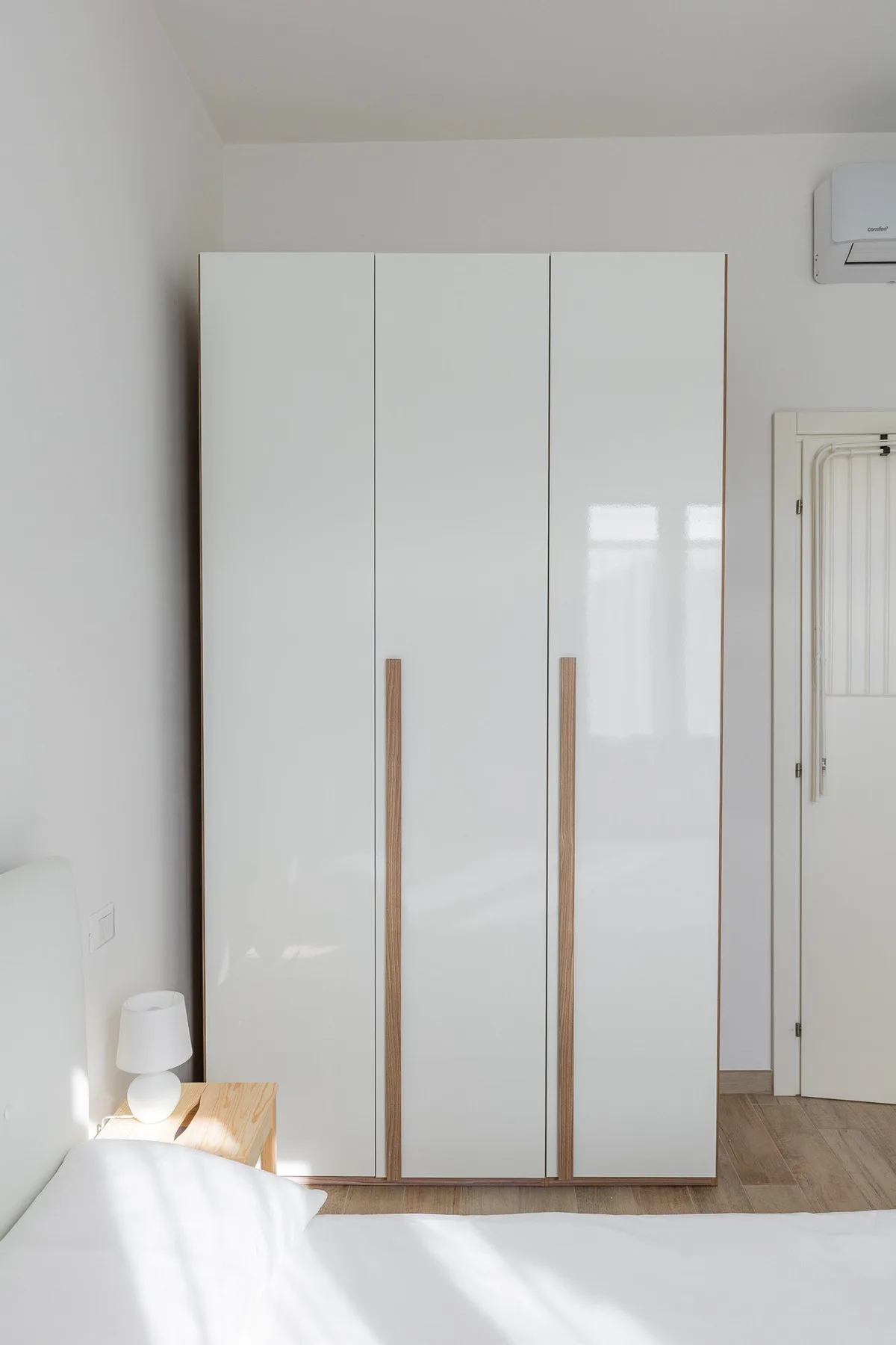 Un dormitorio luminoso con un armario blanco alto junto a la cama.