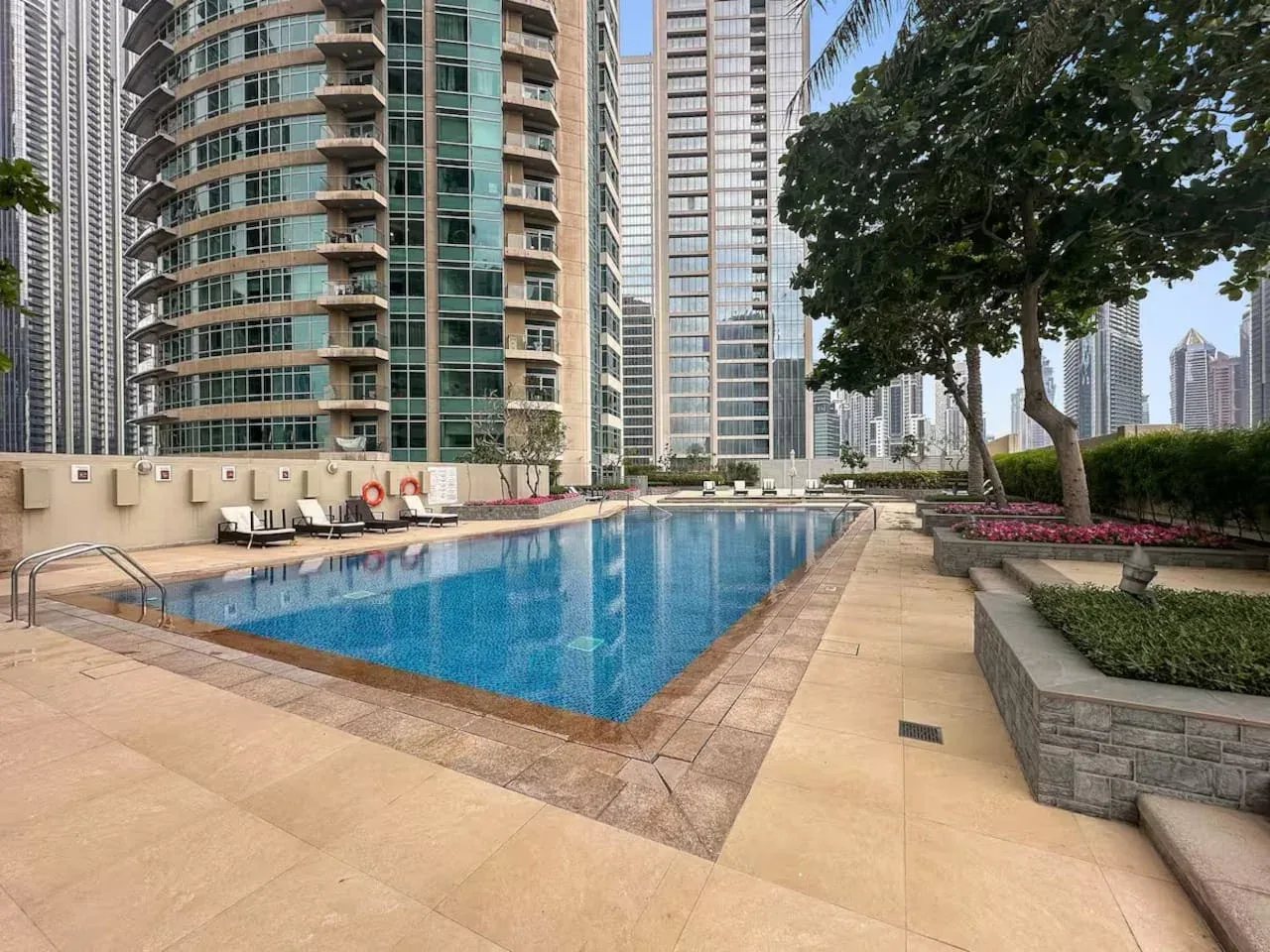 An outdoor swimming pool sits among high-rise buildings, with trees and paved seating areas nearby.