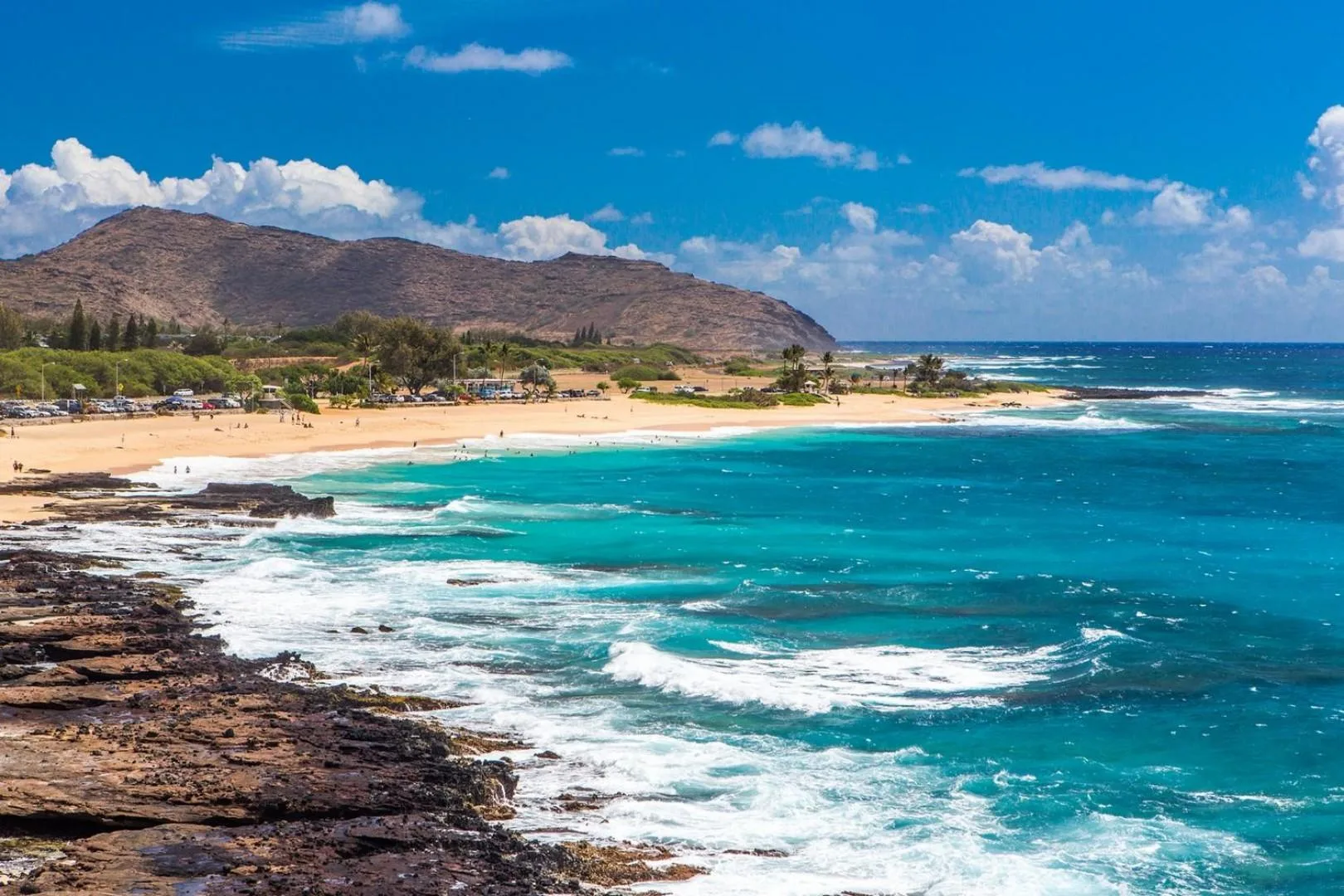 A coastal beach with turquoise waves, rocky shoreline, and mountains in the distance.