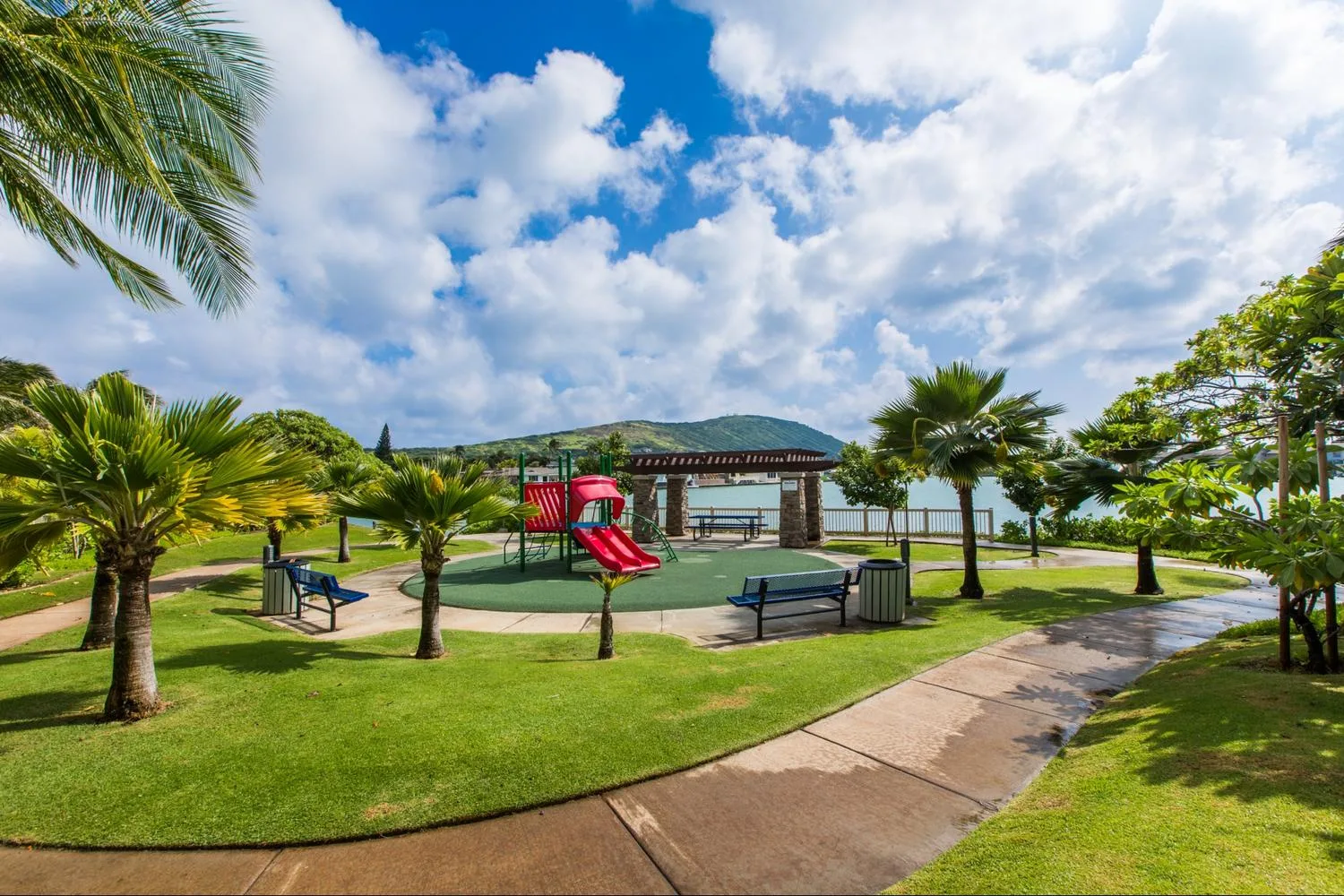 Outdoor playground with a red slide, palm trees, and a paved path.