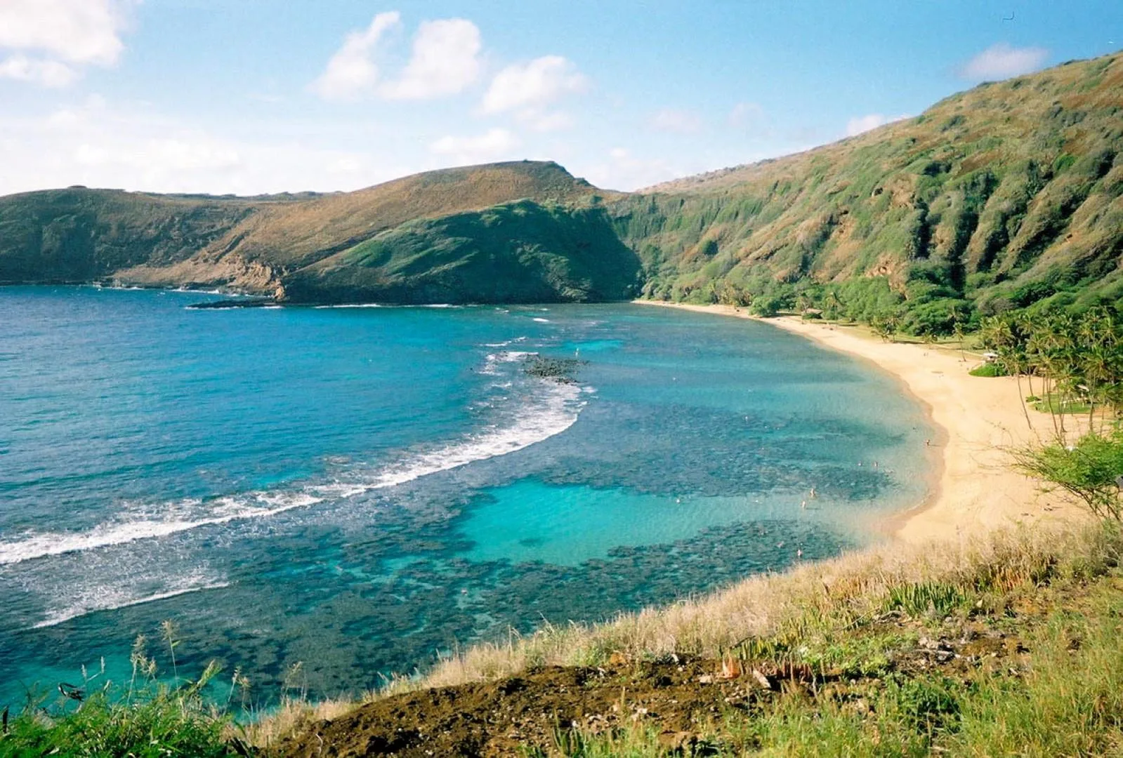 A curved sandy beach borders clear blue water beneath green hills.