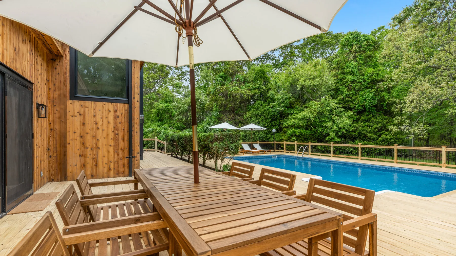 Una mesa de comedor de madera al aire libre está junto a una piscina y árboles verdes.