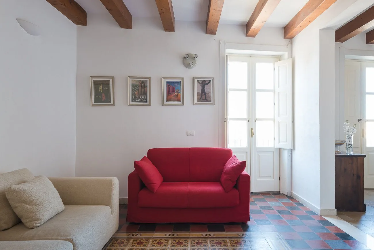 A bright living room with red and beige seating, framed pictures, and exposed ceiling beams.
