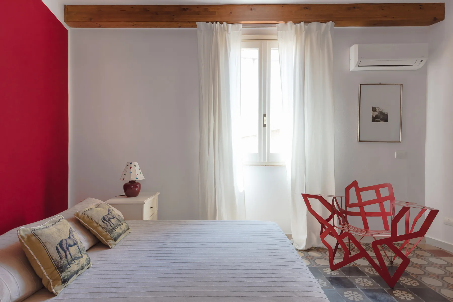 A bedroom with a bed, a bright curtained window, and red chairs.