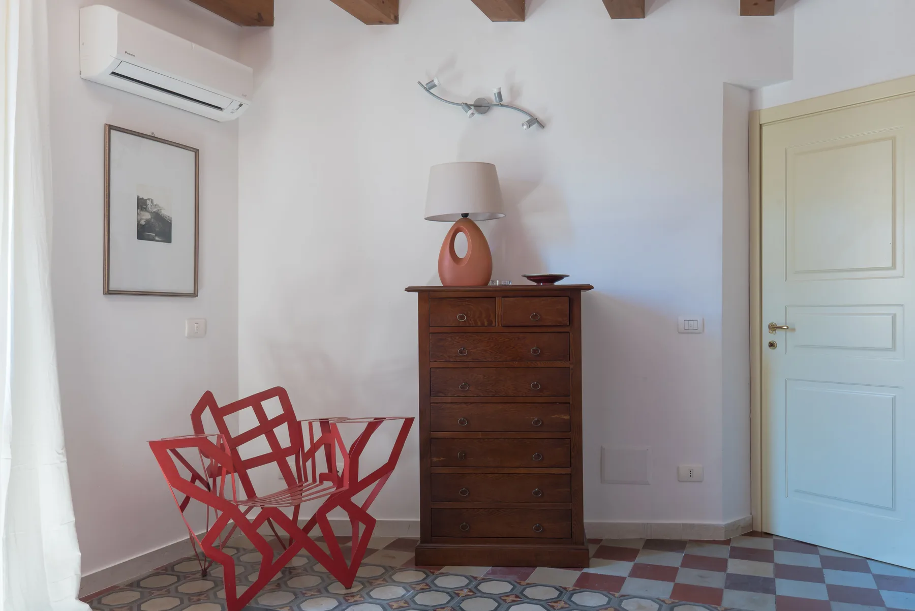 A bright bedroom with a wooden chest of drawers, red seating, and a door.