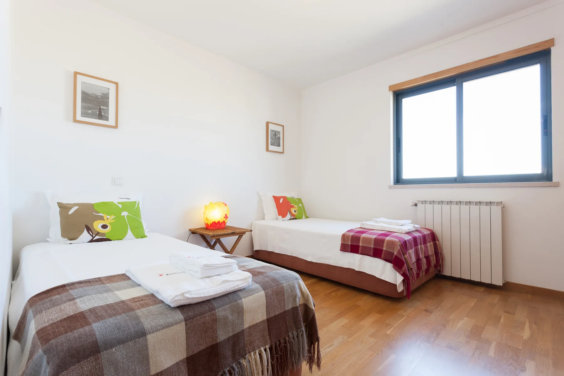 A bright bedroom with two beds, plaid blankets, and a large window.