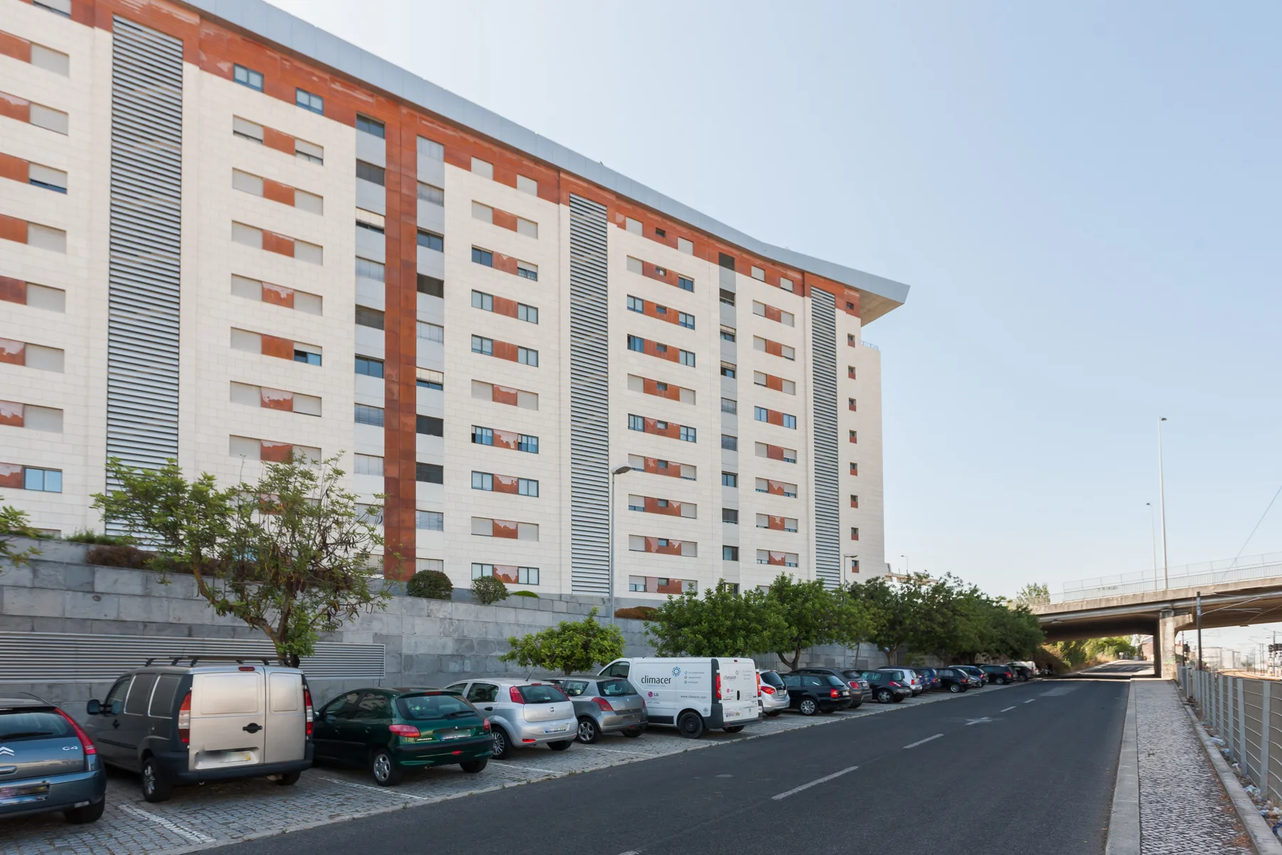 A city street runs beside a large apartment building and parked cars.