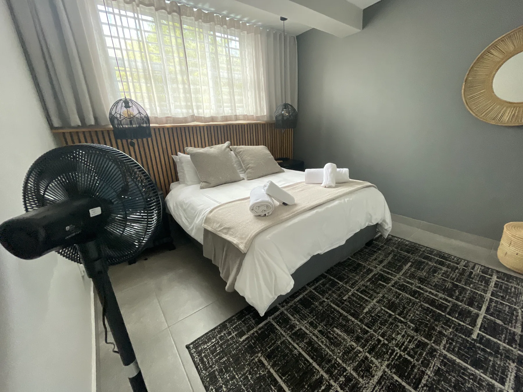 A bedroom with a neatly made bed beside a large curtained window.