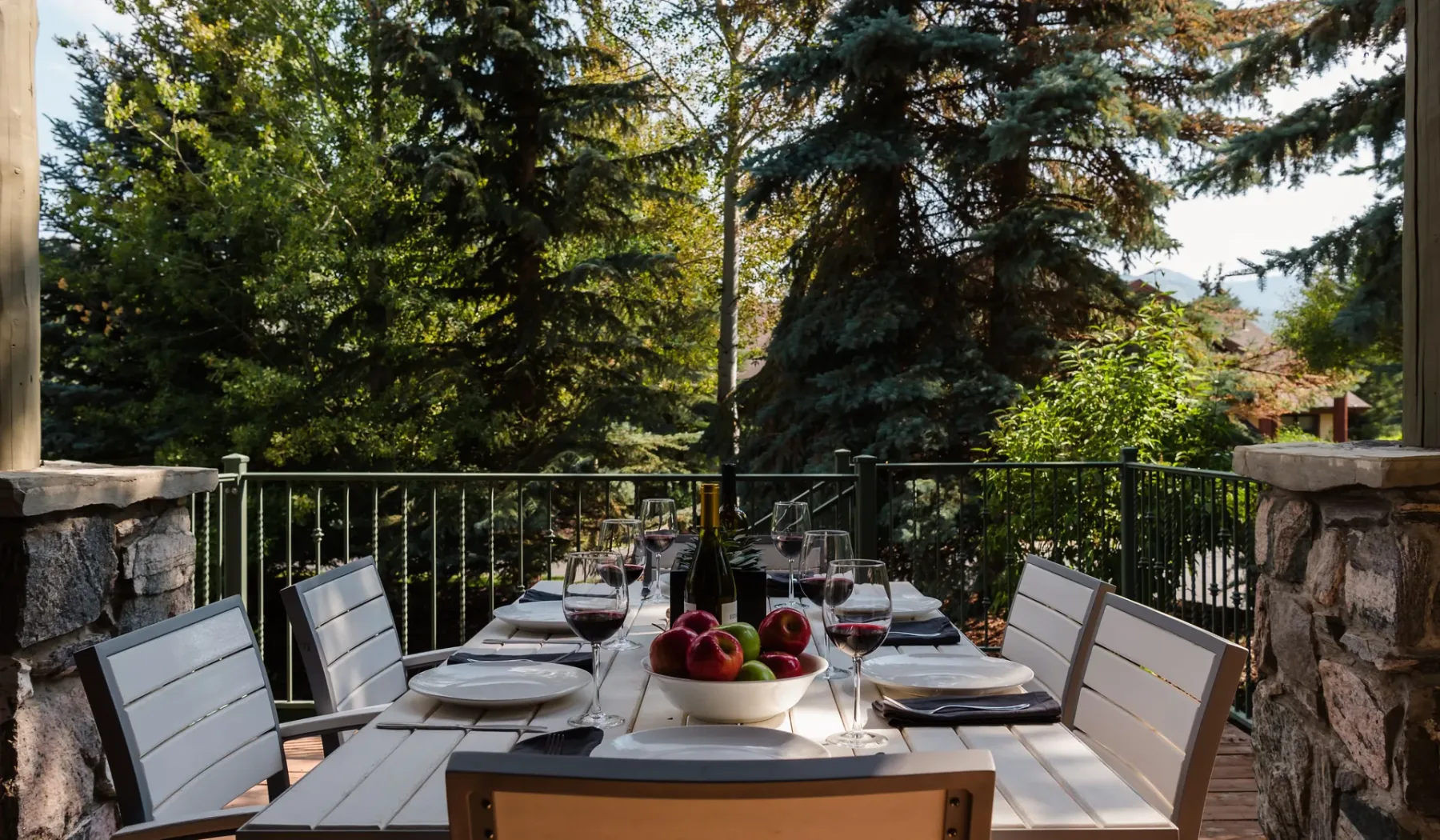 Una mesa de comedor al aire libre está preparada en un patio junto a una barandilla metálica y árboles frondosos.