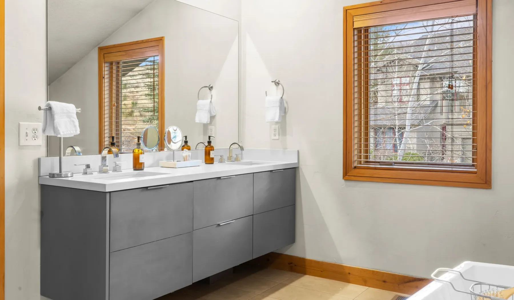 Un baño luminoso con un largo tocador, lavabos sobre la encimera y ventanas con marcos de madera.