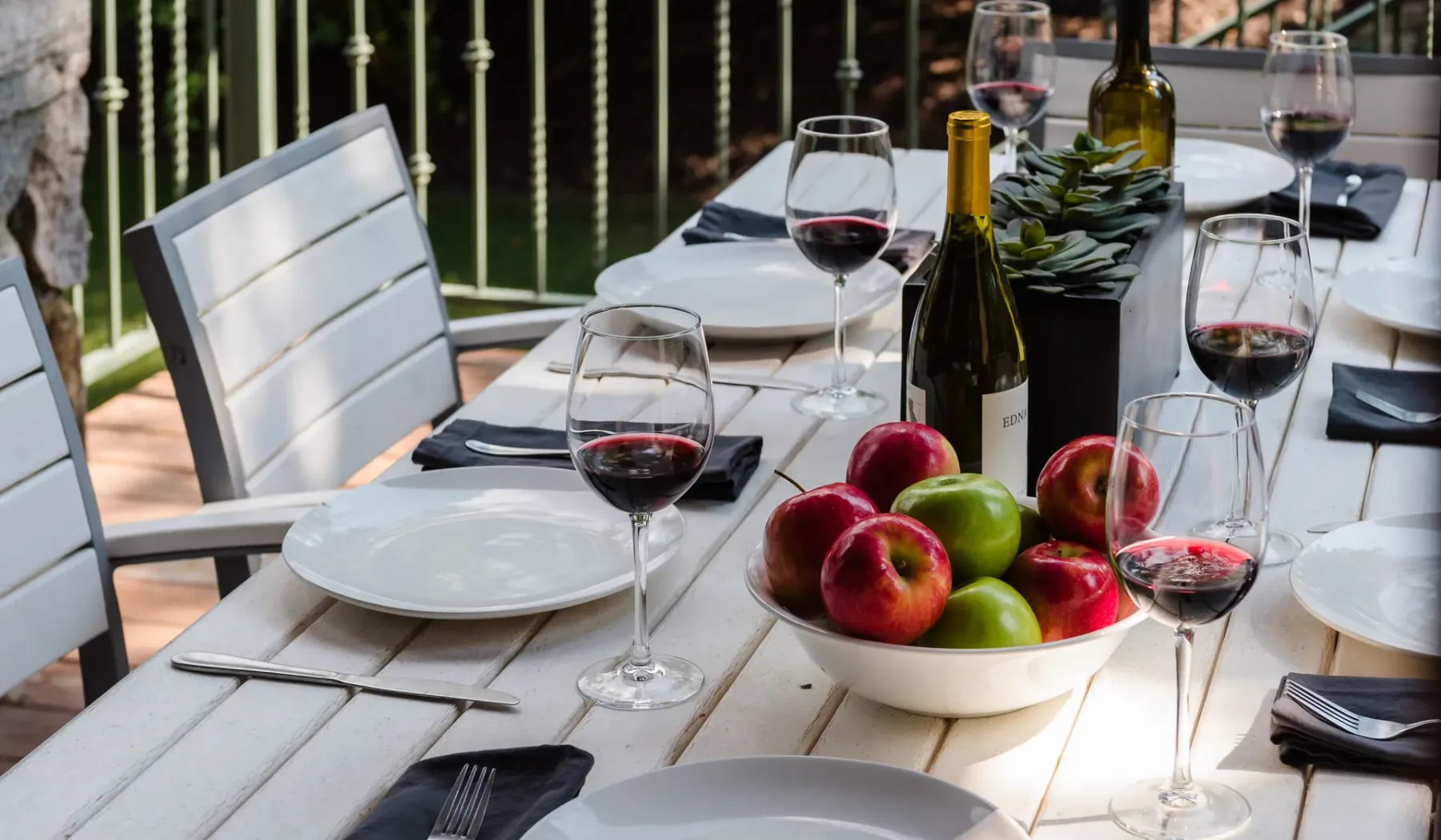 Una mesa de comedor al aire libre está puesta con platos, copas de vino y un cuenco de manzanas.