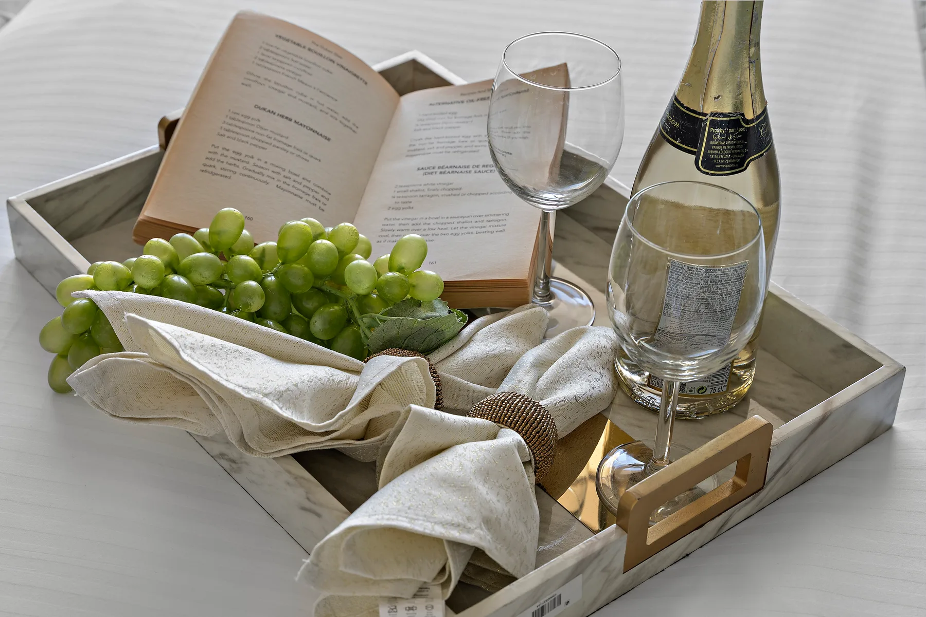 A serving tray holds grapes, folded linens, an open book, and glassware.