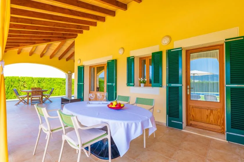 A covered terrace with a dining table and green shutters along the yellow wall.