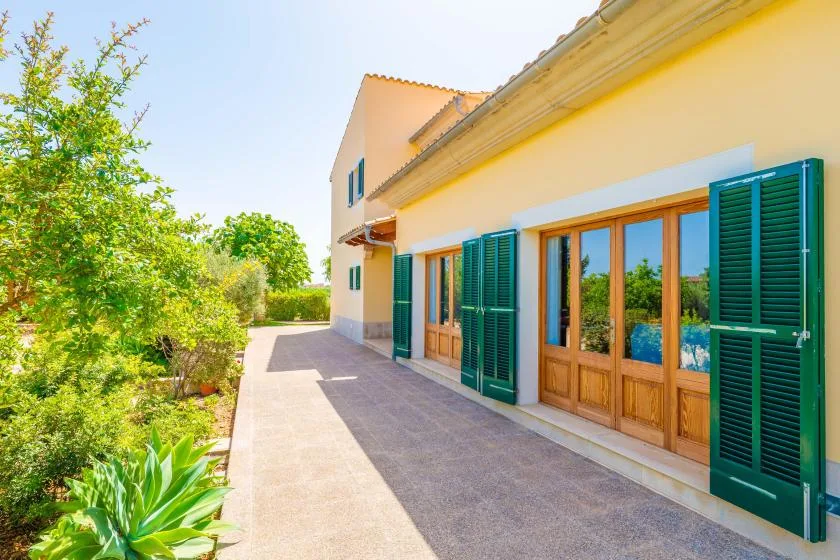 A paved walkway runs beside a yellow house with green shutters and wooden doors.