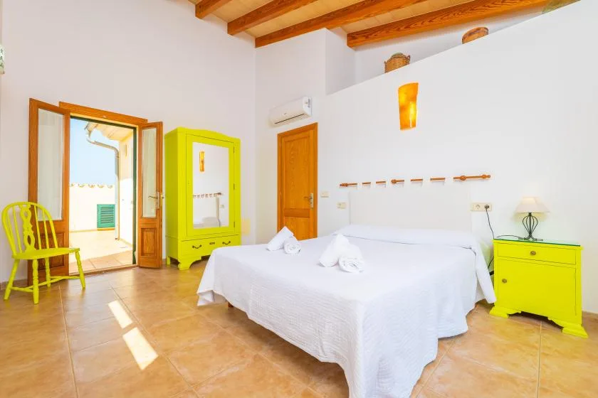 A bright bedroom with a white bed, yellow furnishings, and exposed wooden ceiling beams.
