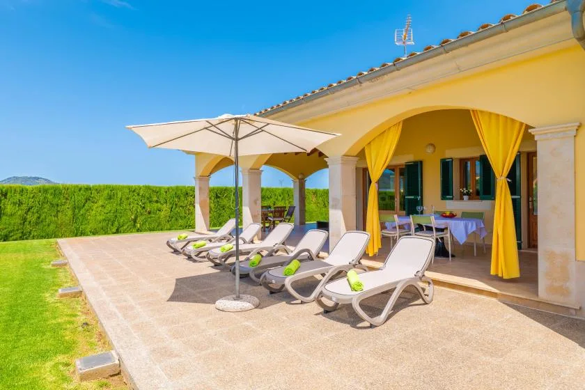 A sunny patio with reclining loungers and a large white umbrella beside a yellow-curtained veranda.