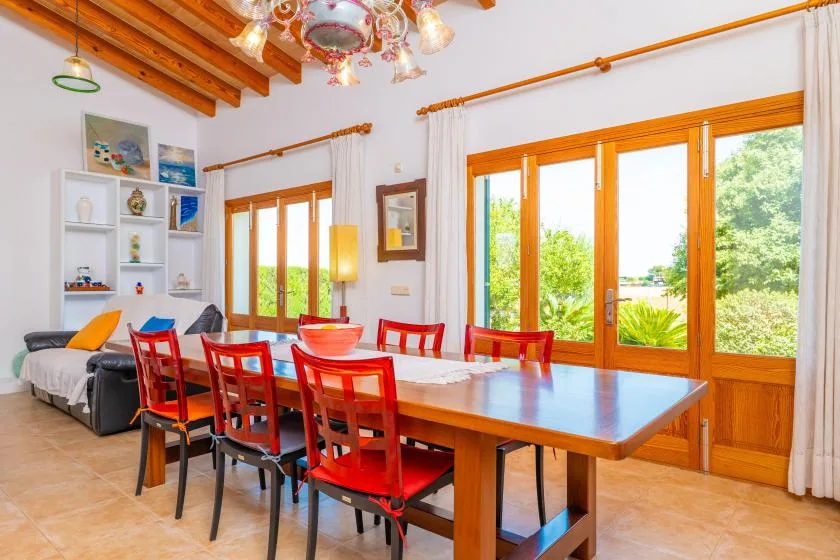 A dining area with a long wooden table and red chairs beside large windows.