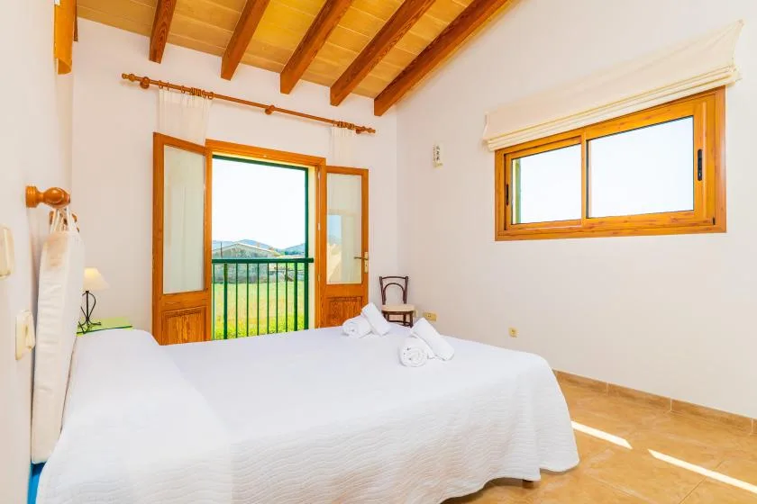 A bright bedroom with a white bed, exposed wooden ceiling beams, and windows.