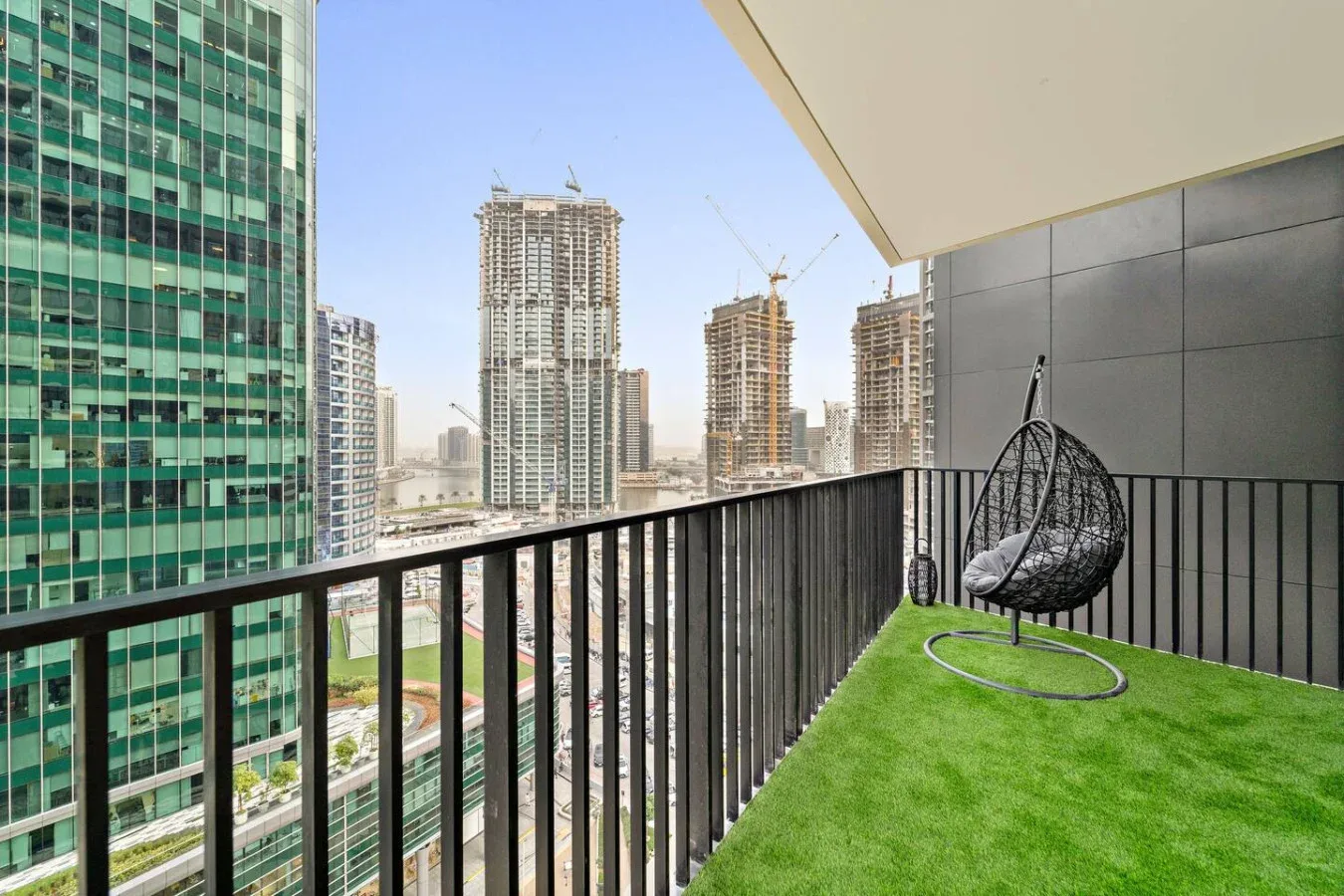 Un balcón con césped artificial y una silla colgante da a edificios altos cercanos.