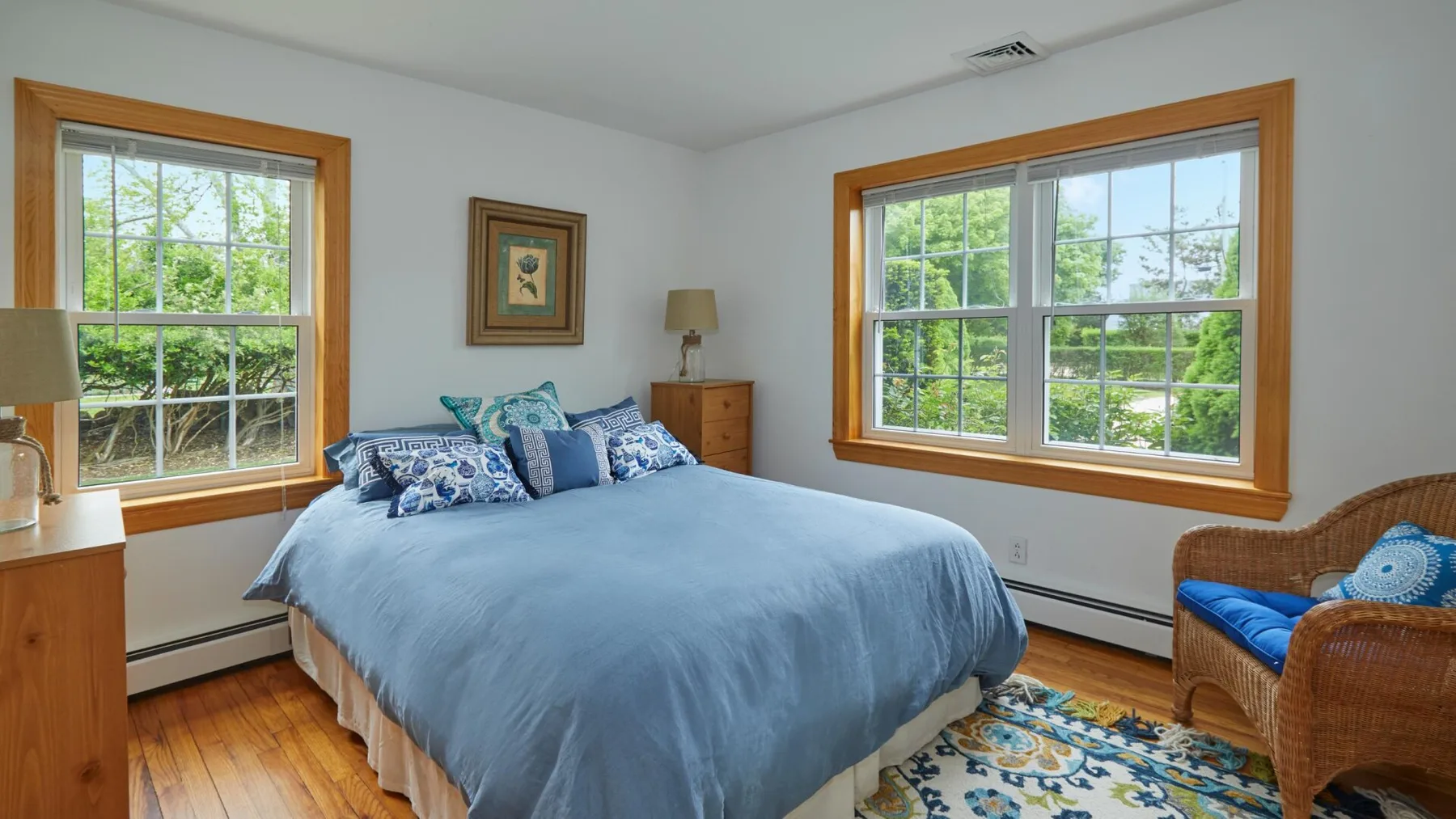 Un dormitorio con una cama, ventanas con molduras de madera y una silla de mimbre.