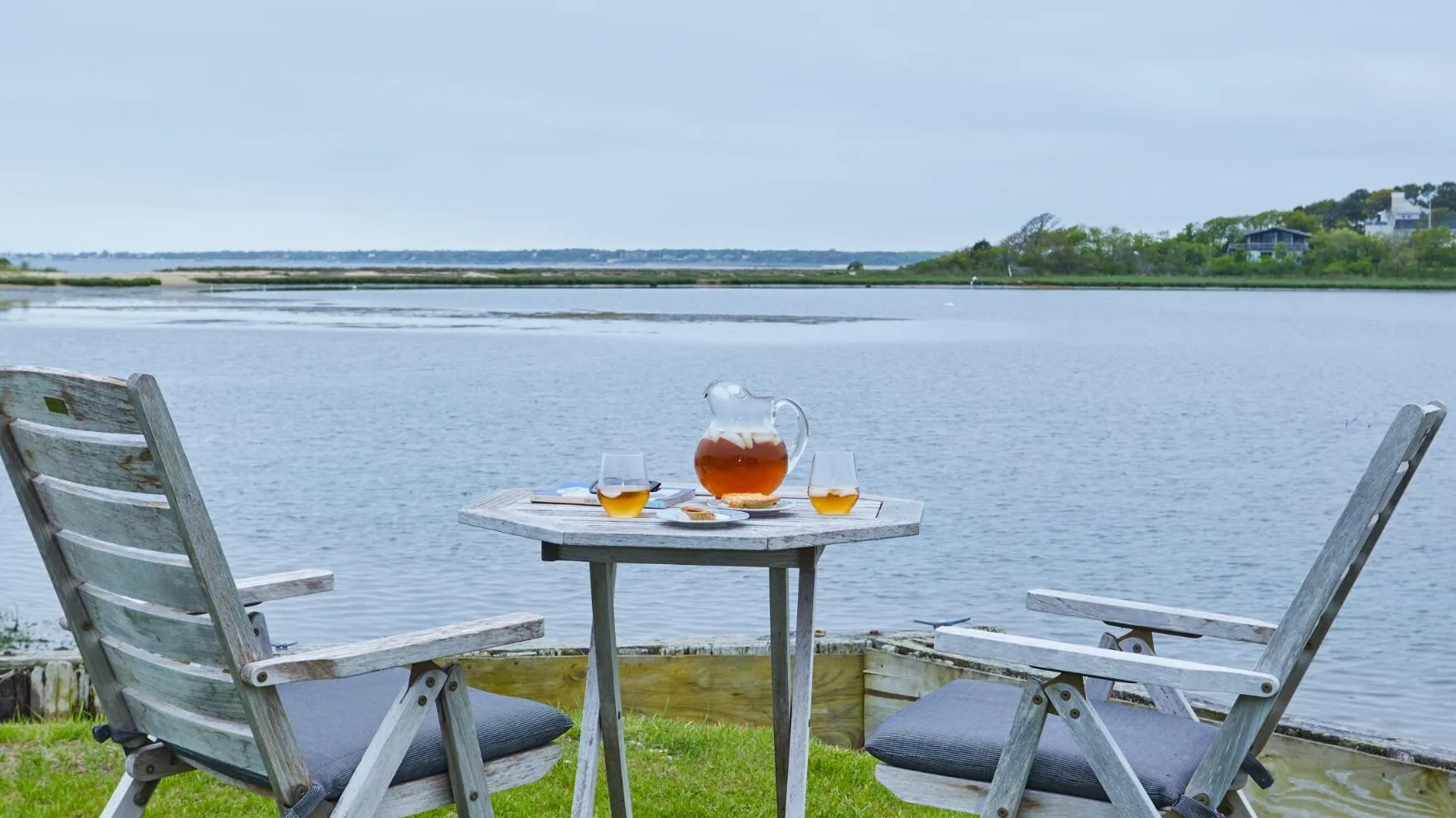 Dos sillas al aire libre y una mesa pequeña con bebidas miran hacia una masa de agua tranquila.