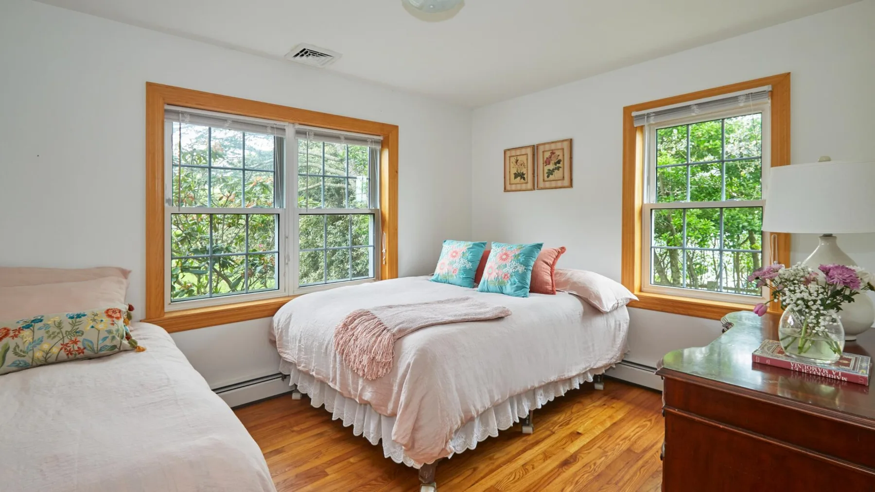 Un dormitorio luminoso con una cama entre dos ventanas y una cómoda de madera.