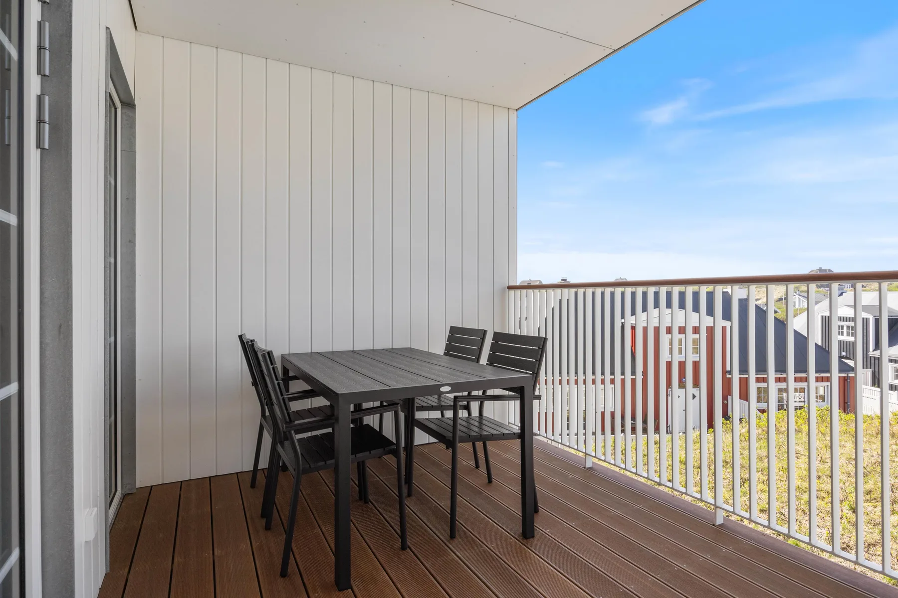 Un balcón cubierto tiene una mesa de exterior oscura y asientos junto a una barandilla blanca.