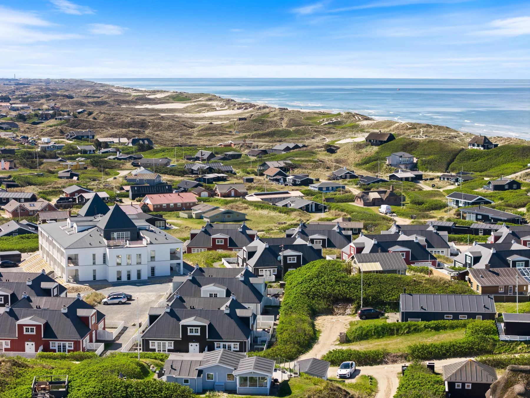Vista aérea de un barrio costero con casas, dunas y el mar al fondo.