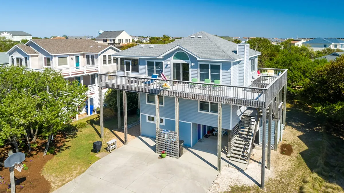 A raised blue beach house with a broad wooden deck and exterior stairs.