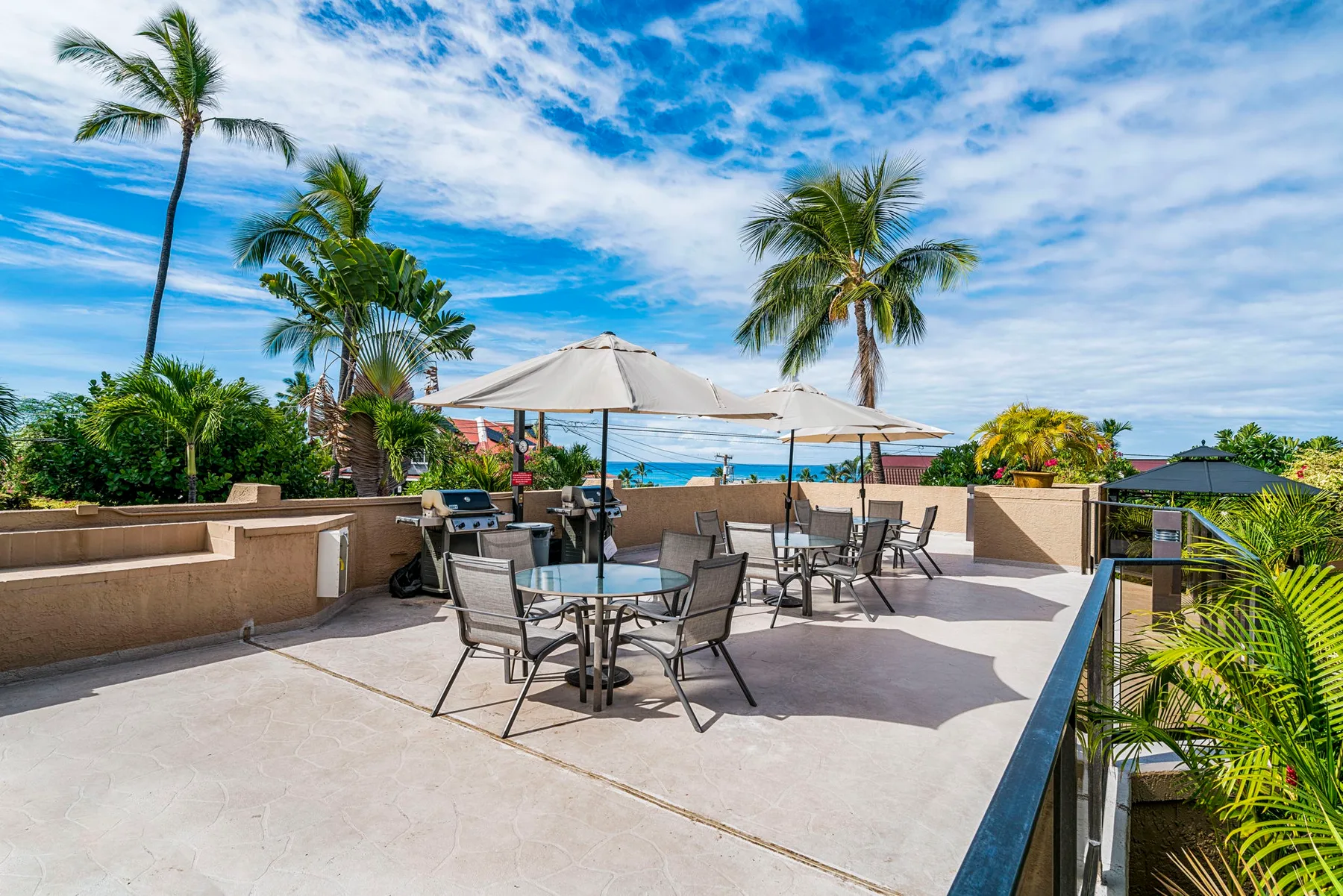 Un patio al aire libre tiene mesas bajo sombrillas, con palmeras y vistas al mar.