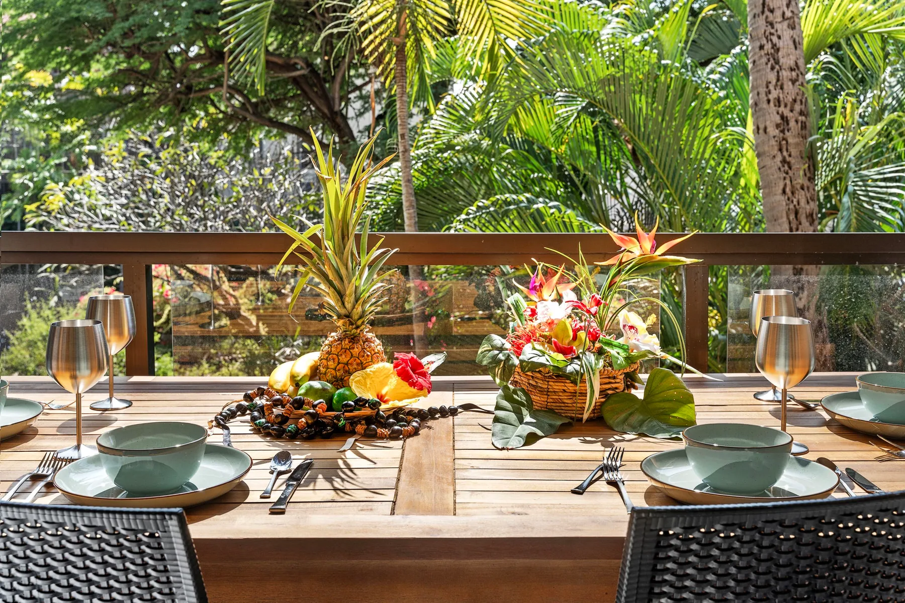 Una mesa de comedor al aire libre está puesta con fruta, flores, platos y copas entre vegetación tropical.