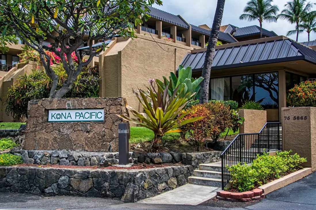 Entrada de un edificio ajardinada, con un letrero de piedra, escalones y plantas tropicales.