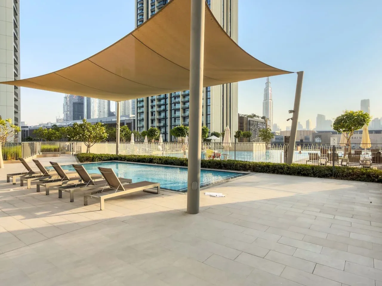 An outdoor pool and lounge area sits beneath a large shade canopy, with city buildings beyond.