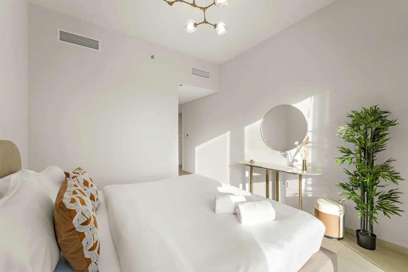 A bright bedroom with white bedding, a round mirror, and a potted plant.