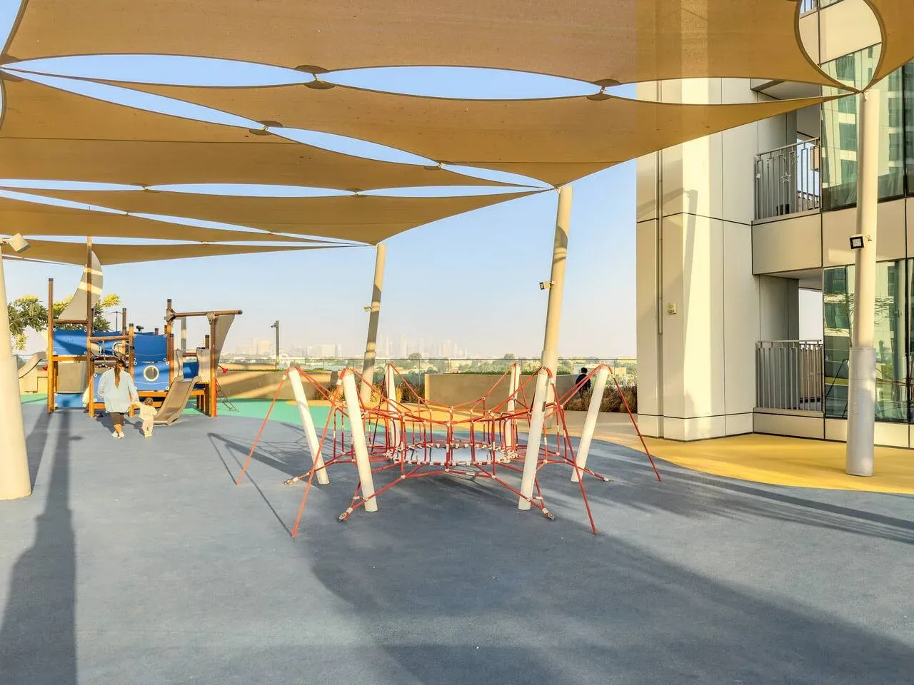 An outdoor playground sits beneath broad shade canopies beside a modern building.