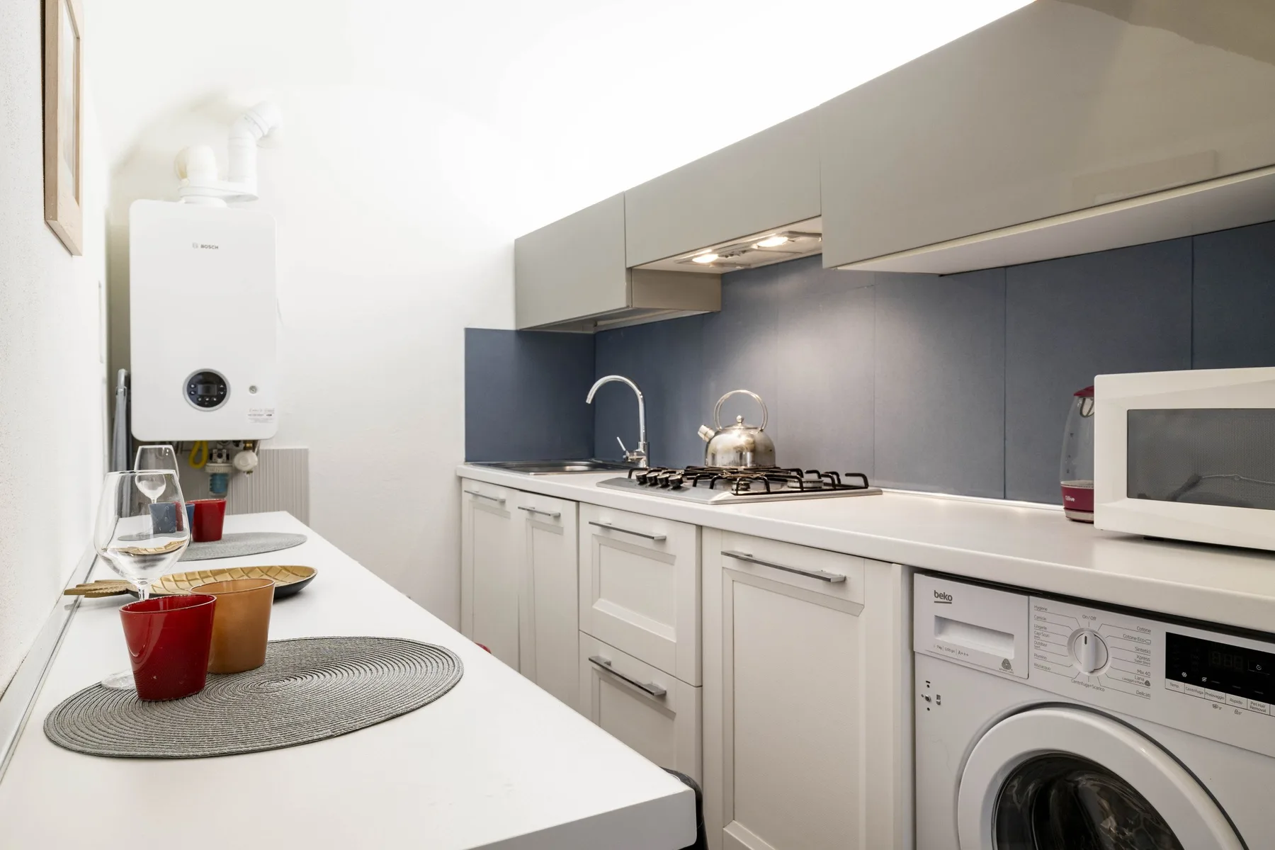 Una cocina compacta con armarios blancos, una placa de cocina y una lavadora.