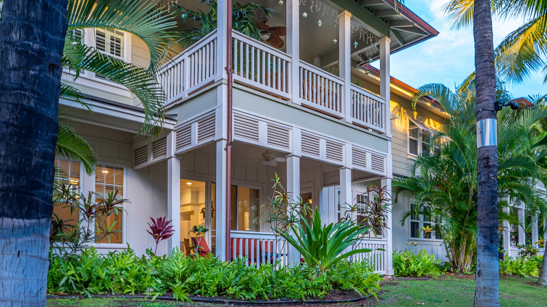 Una casa blanca de dos pisos con porches rodeada de plantas tropicales y palmeras.