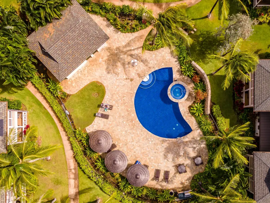 Vista aérea de una piscina curva rodeada por un patio pavimentado y vegetación tropical.