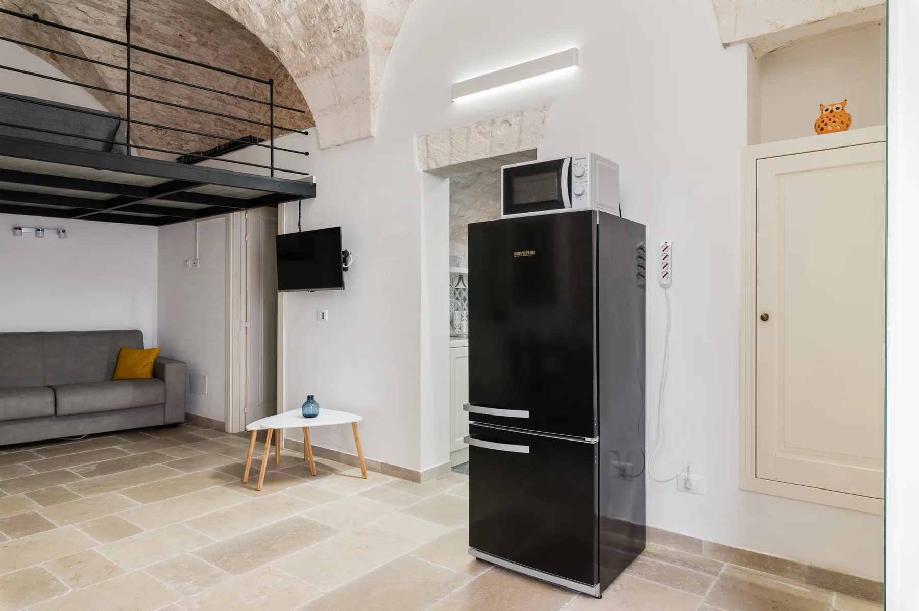 Una pequeña zona de estar con sofá gris, mesa pequeña y refrigerador negro.