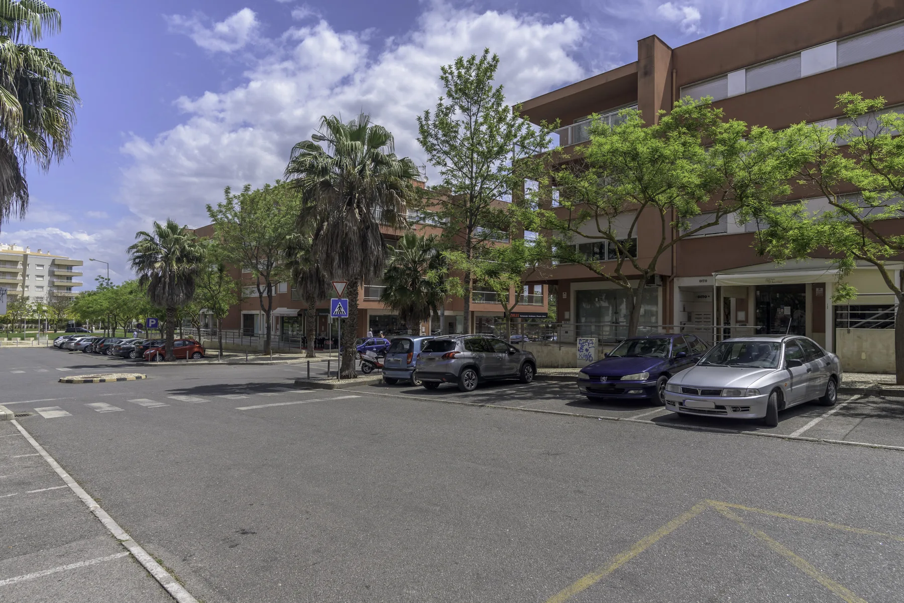 Una vista de la calle muestra un edificio moderno detrás de coches aparcados y árboles frondosos.