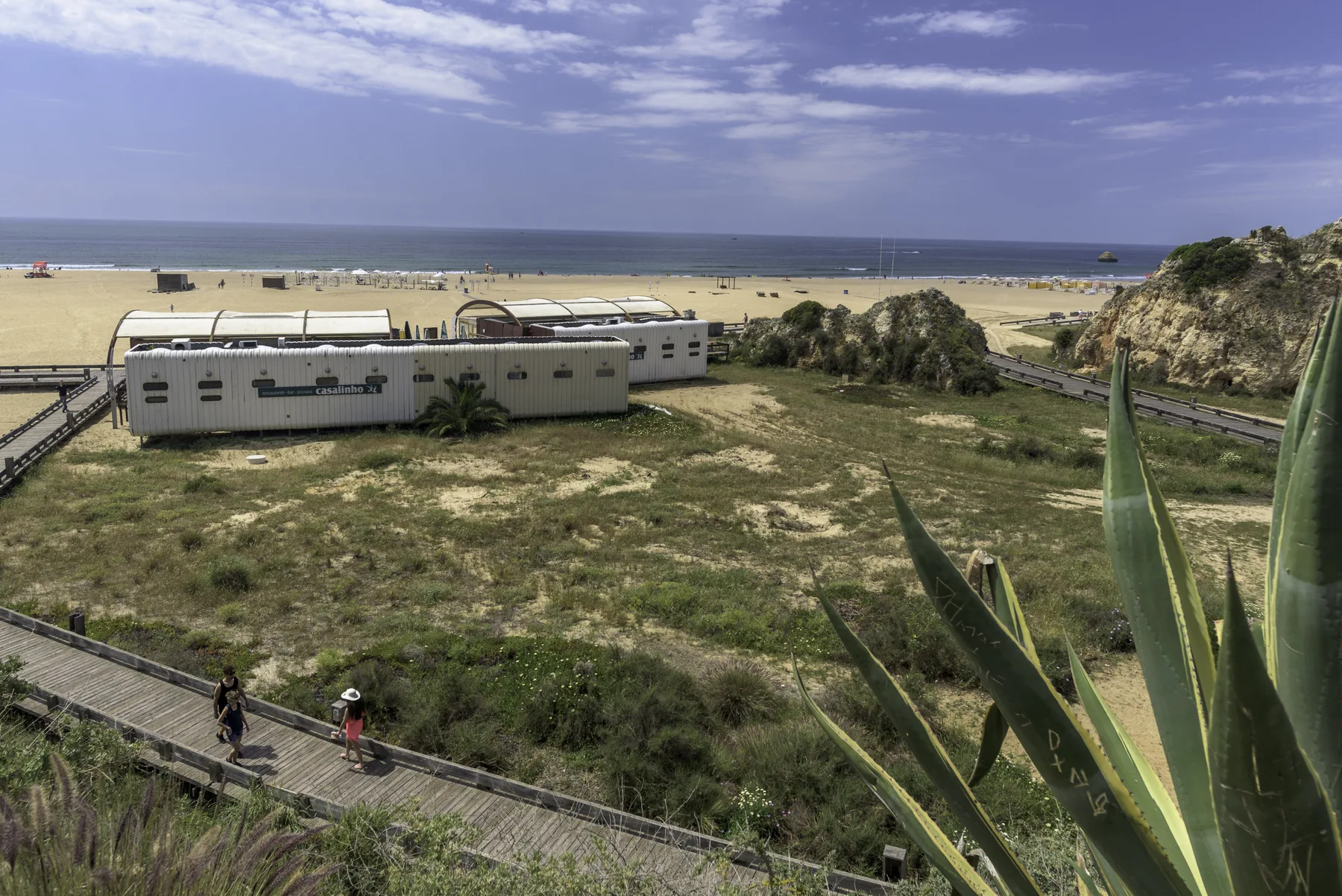 Paisaje costero con una playa de arena, edificios bajos y una pasarela de madera.