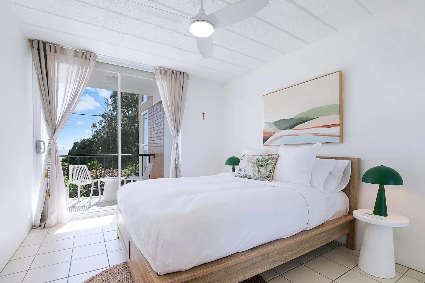 Un dormitorio luminoso con una cama, ventilador de techo y puertas de vidrio que dan a un balcón.