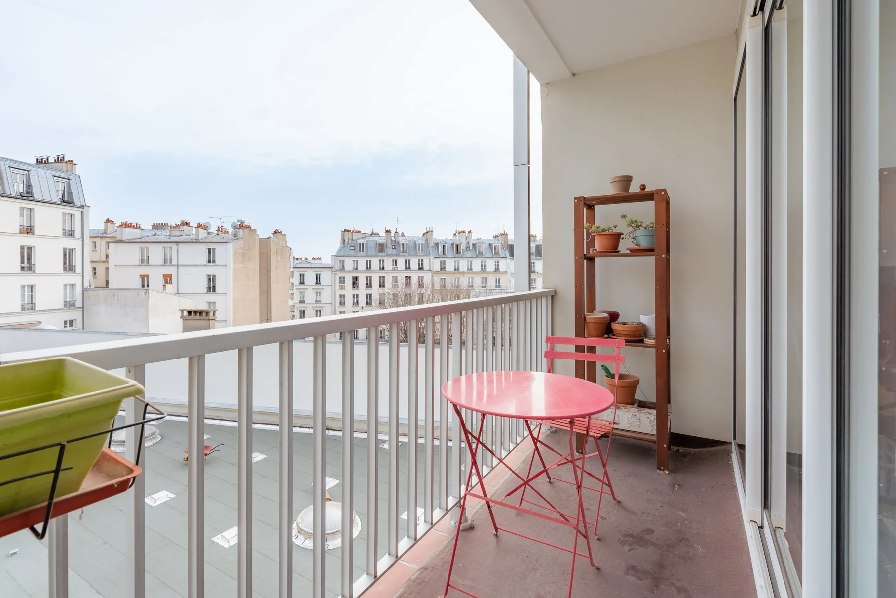 A balcony with a red table and chairs overlooking nearby buildings.