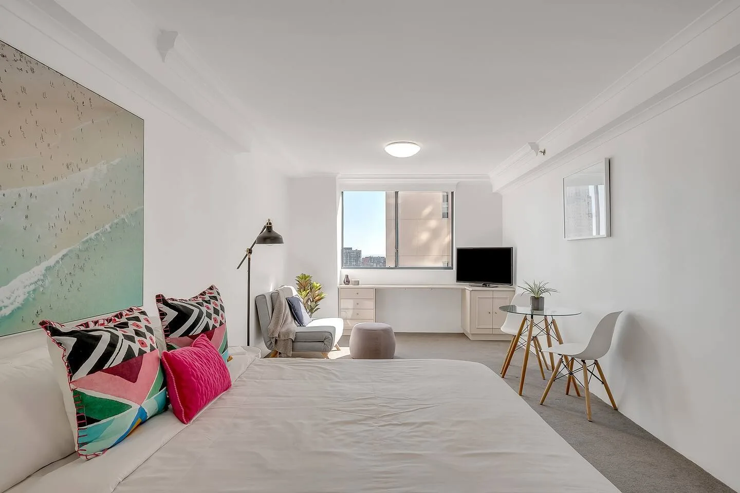 A bright studio bedroom with a bed, seating area, and window overlooking nearby buildings.