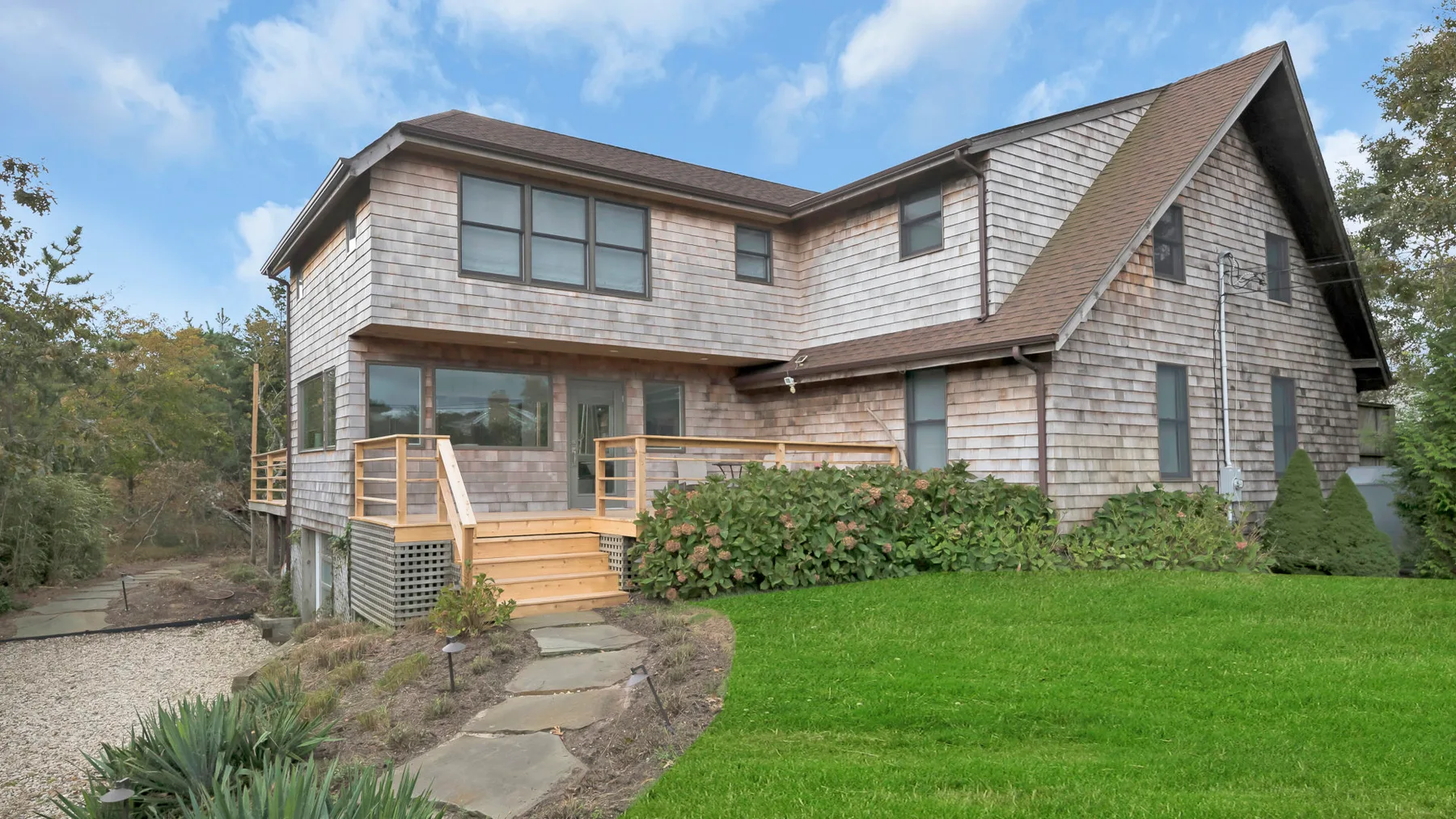 A shingled house exterior with a wooden deck, garden shrubs, and a green lawn.