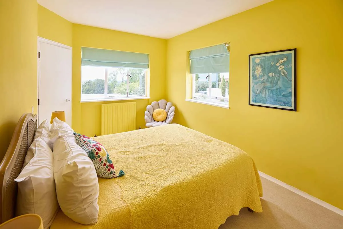 Un dormitorio luminoso con una colcha amarilla y dos ventanas.