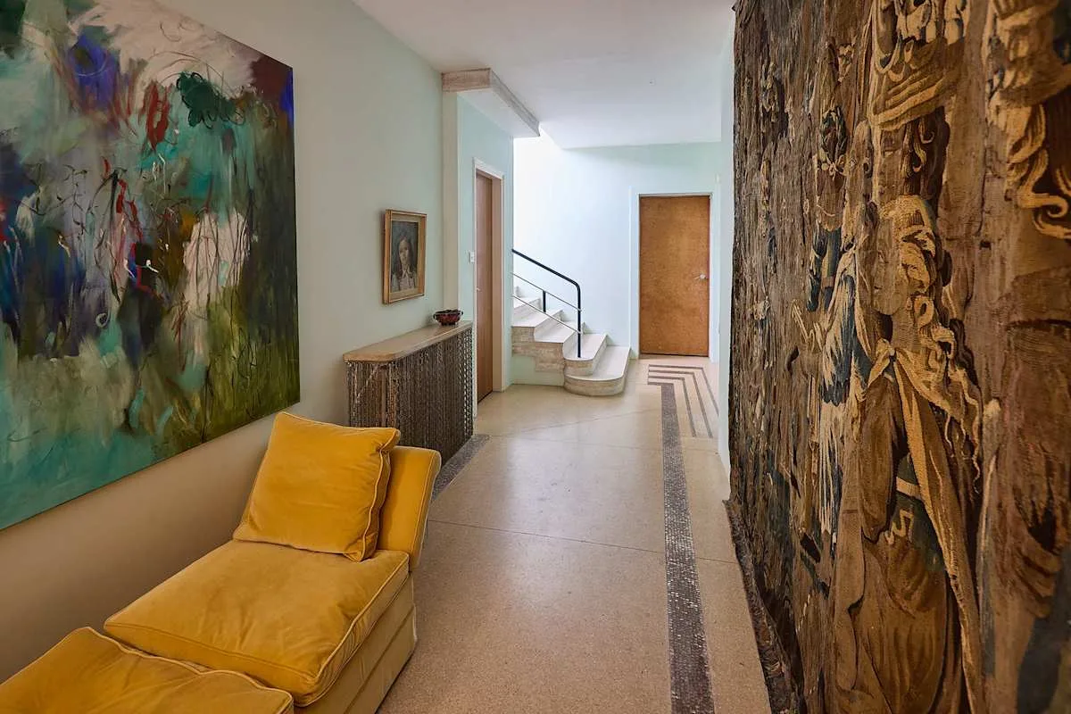 Un pasillo luminoso presenta obras coloridas y una chaise longue amarillo mostaza.