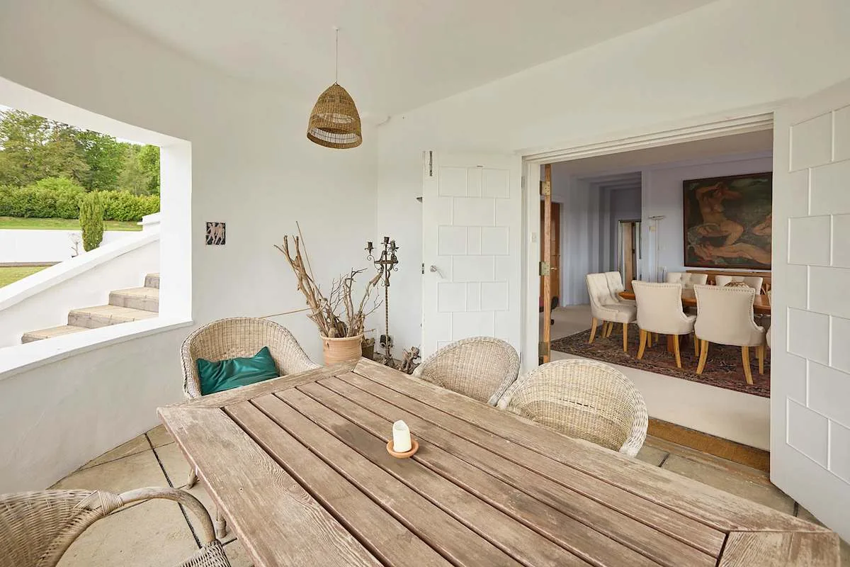 Un comedor luminoso da a una terraza cubierta con asientos de mimbre y una mesa de madera.