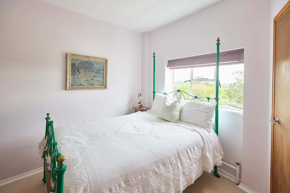 Un dormitorio luminoso con una cama blanca y una ventana con vistas a la vegetación.
