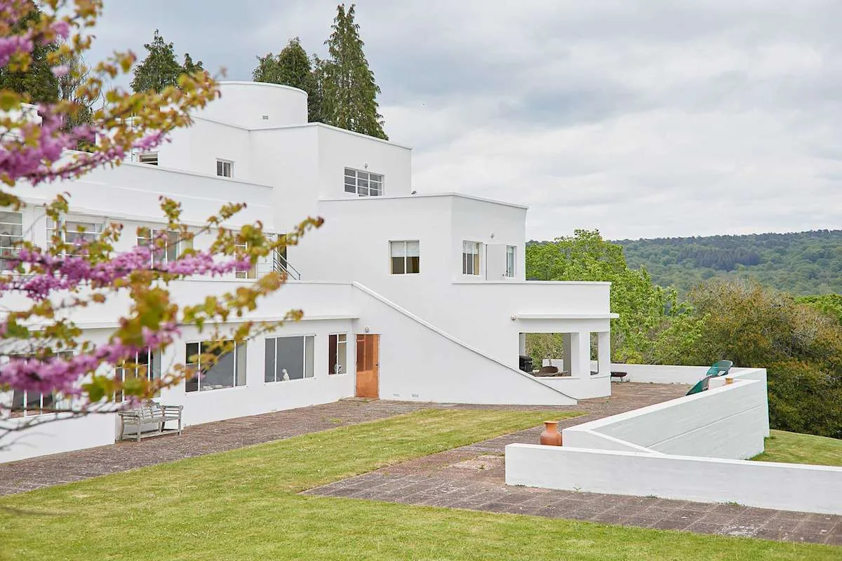 Una casa blanca moderna da a un césped y colinas boscosas a lo lejos.